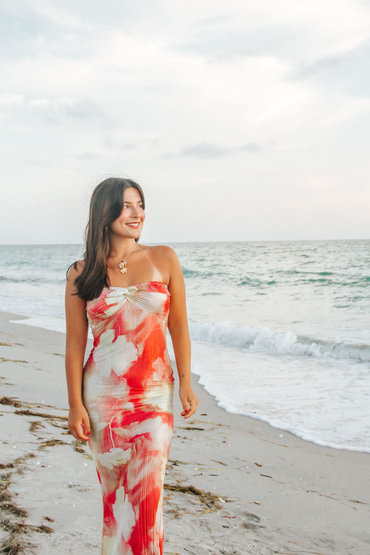 Woman in red floral maxi dress smiling on a sandy beach with ocean waves in the background.