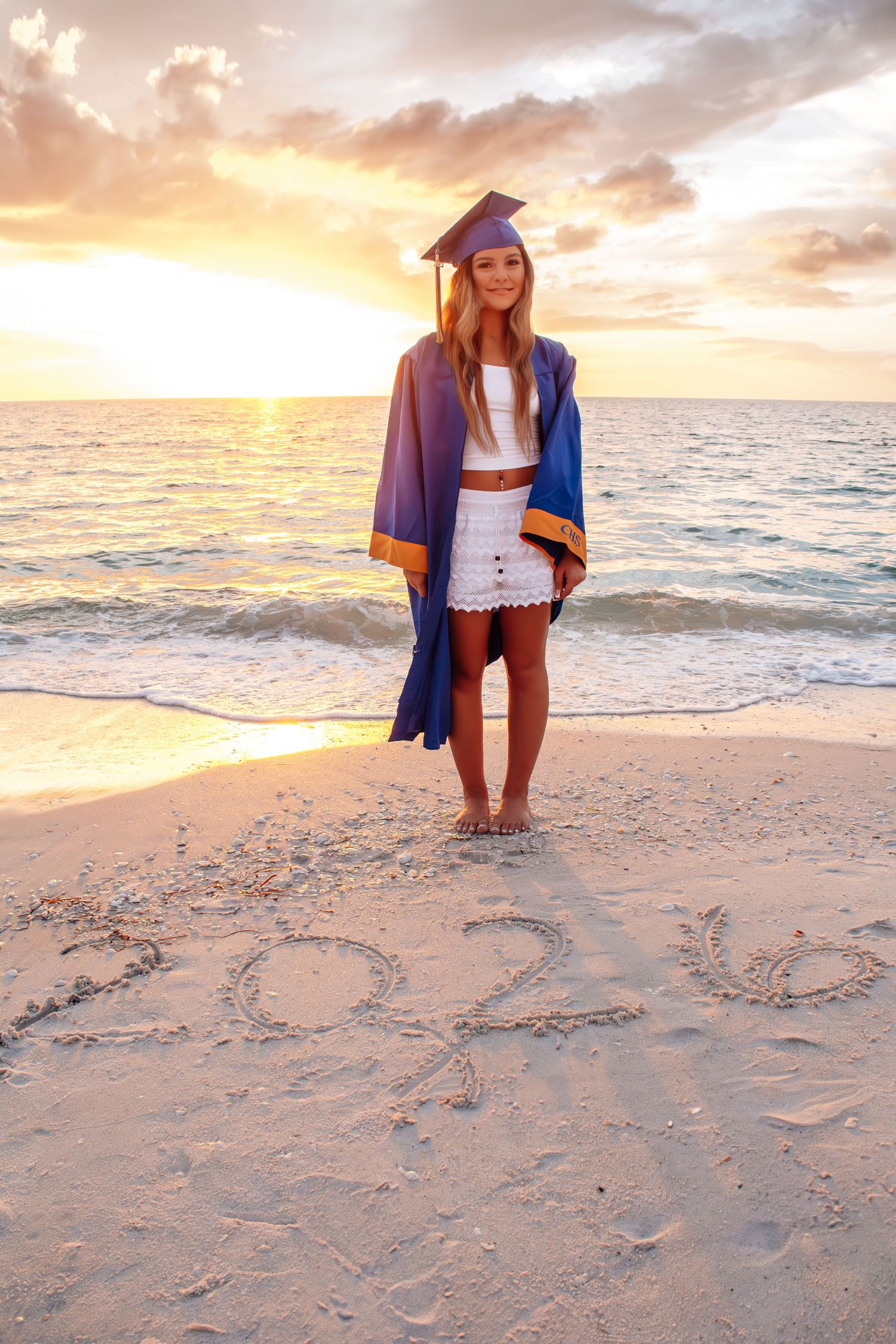 Graduate in cap and gown stands on beach at sunset with 2024 written in sand.