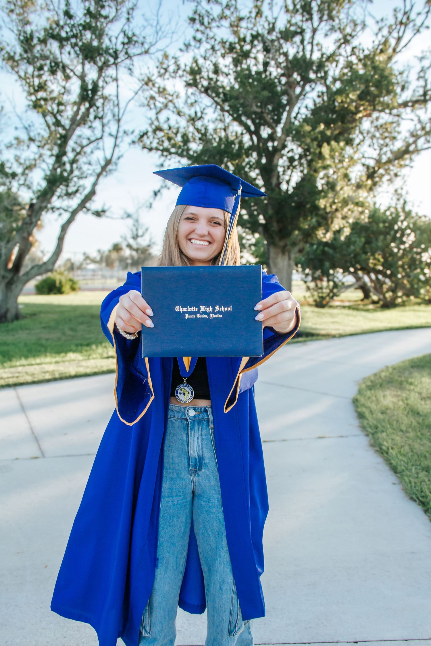 Smiling female graduate in blue cap and gown holding Charlotte High School diploma outdoors.