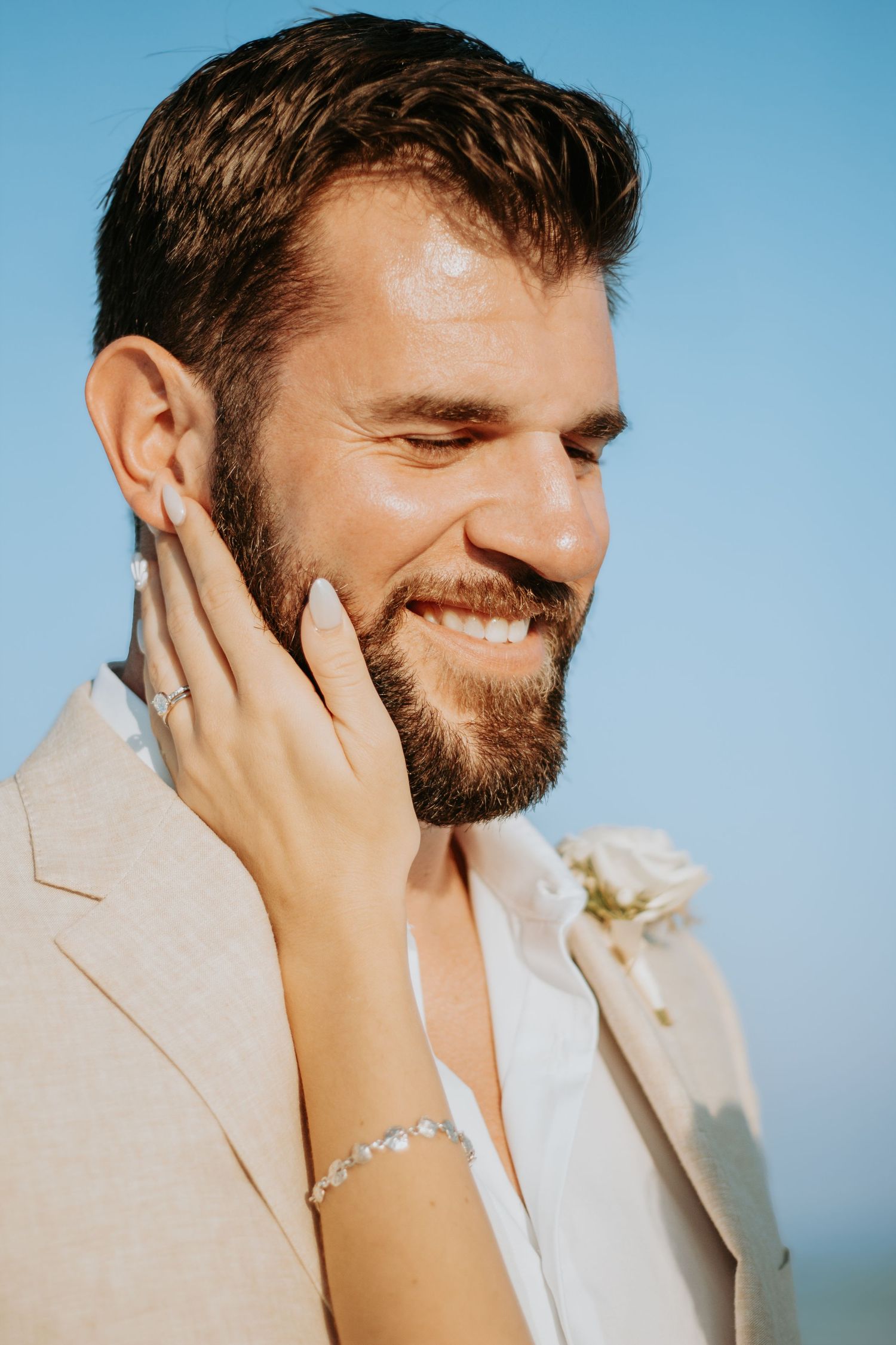 Smiling groom in beige suit touched tenderly on cheek by bride wearing diamond bracelet against blue sky.