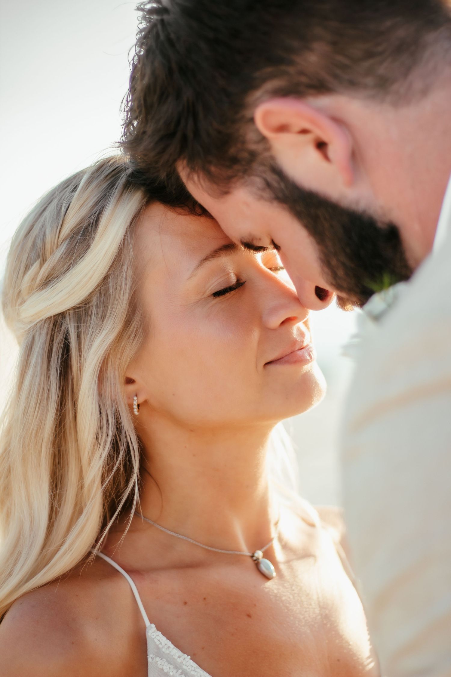 A bearded groom tenderly kisses the forehead of a blonde bride wearing a delicate necklace outdoors.