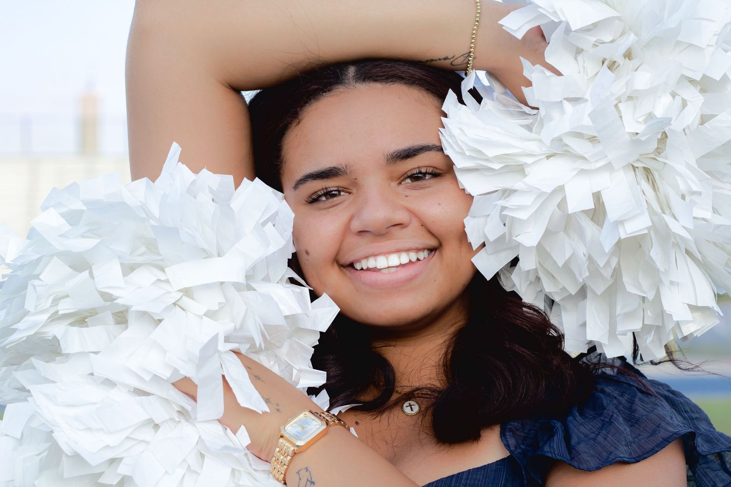 Smiling cheerleader holding white pom-poms, wearing a gold watch and blue outfit, posing outdoors.