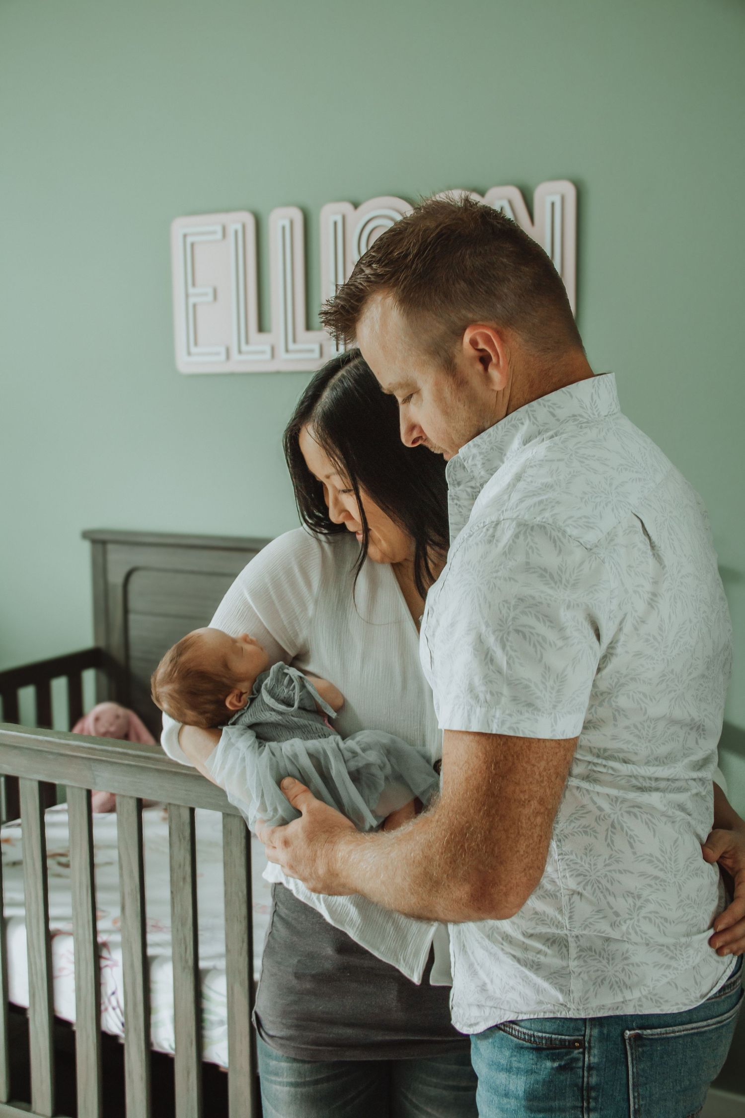 New parents admire their newborn baby in a mint green nursery with the name Ellison on the wall.