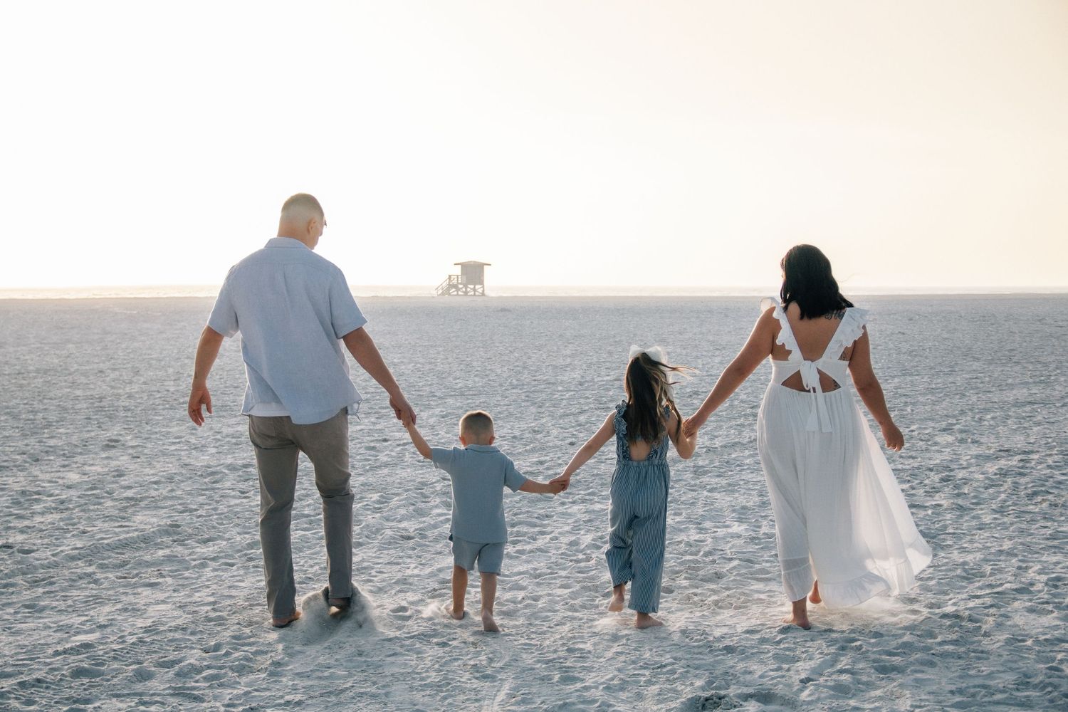 Family of four holding hands walking on a sandy beach toward a lifeguard tower at sunset.