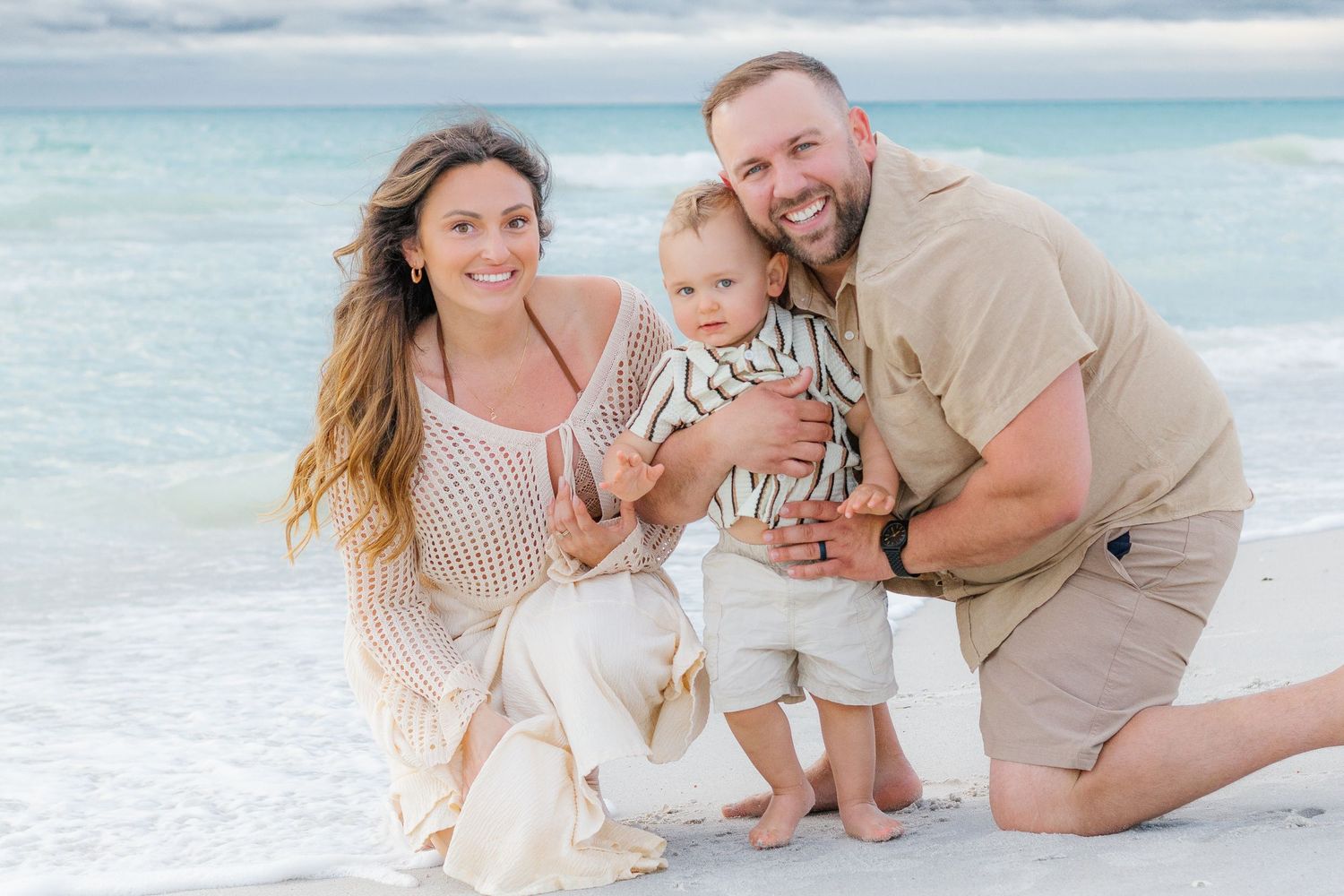 Happy family of three posing on a white sandy beach, parents smiling with their toddler on a sunny day.