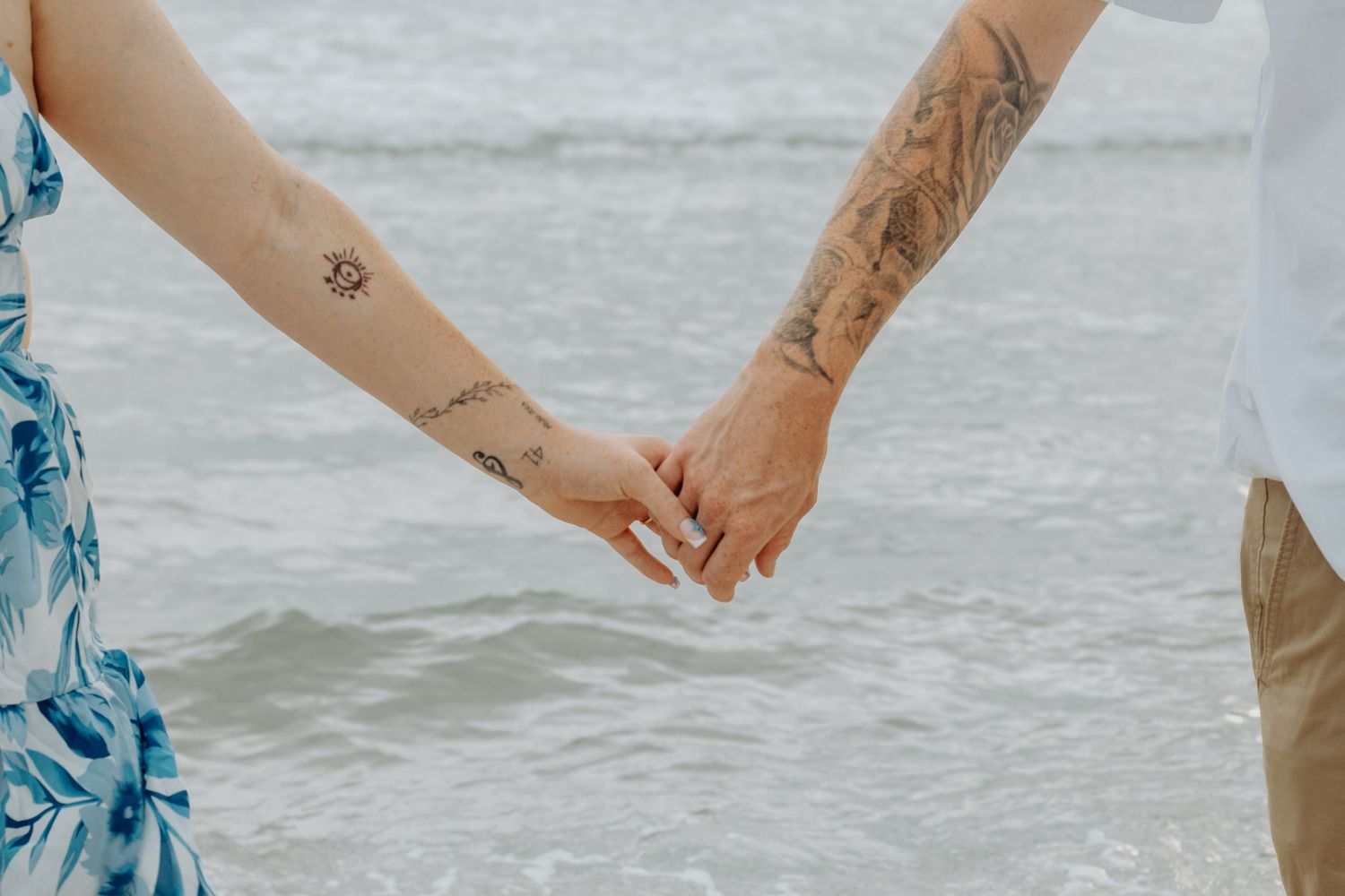 Two people with tattoos holding hands at the beach with ocean waves in the background.