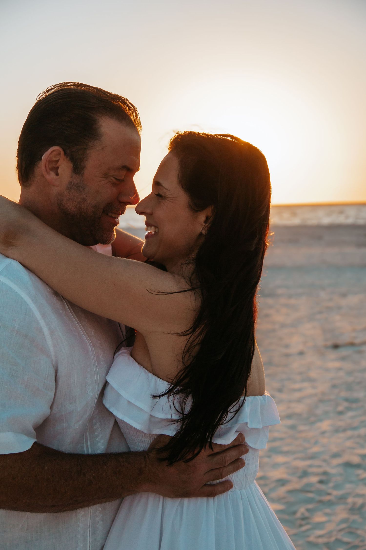 Couple embracing and smiling at sunset on a beach, woman in white off-shoulder dress, romantic golden hour.