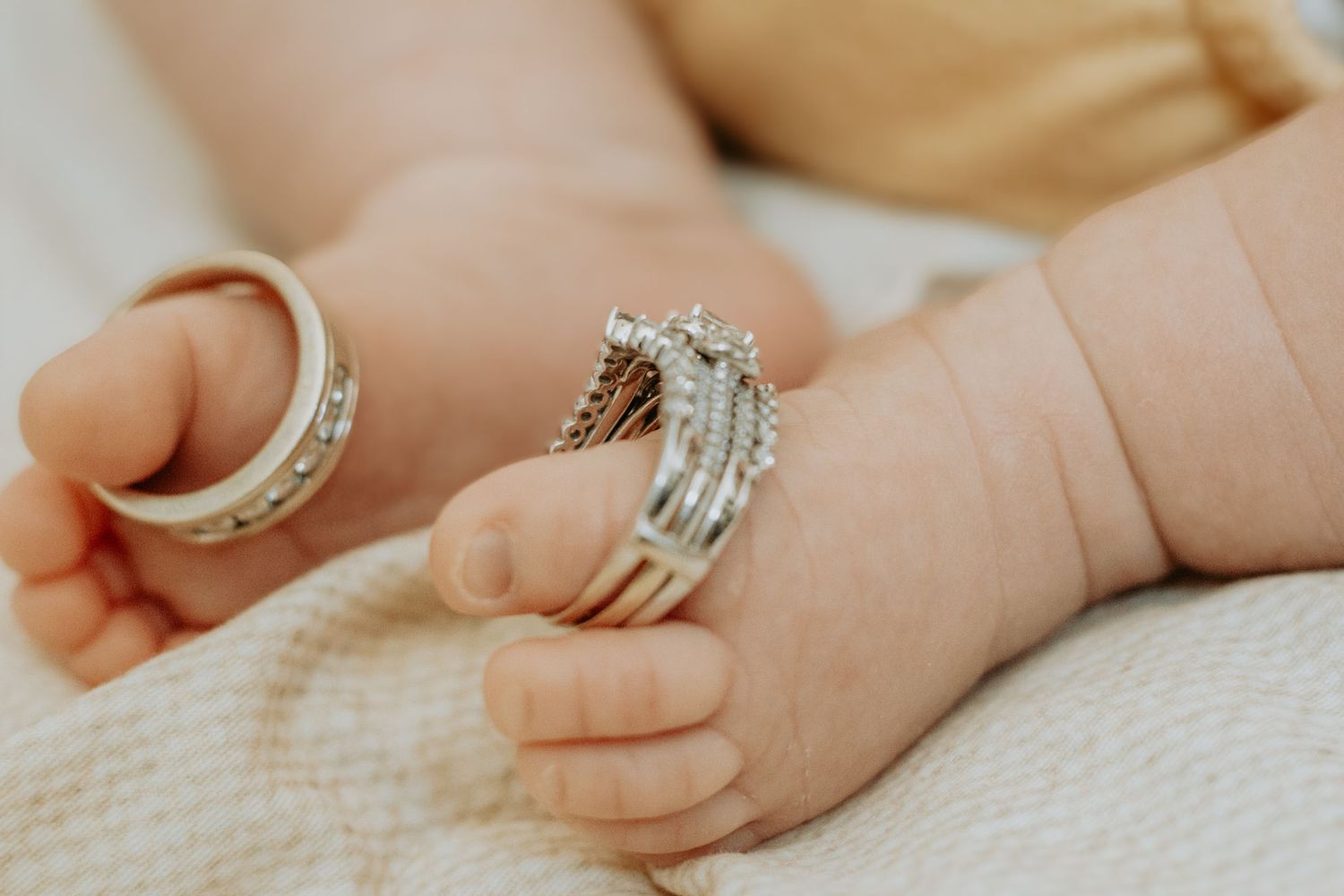 Close-up of a newborn baby's tiny hands adorned with sparkling diamond wedding rings and a gold band.