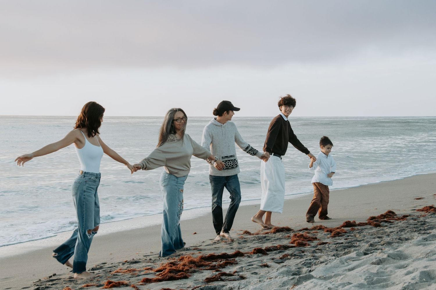 A group of friends in jeans holding hands while walking along a misty beach shoreline.