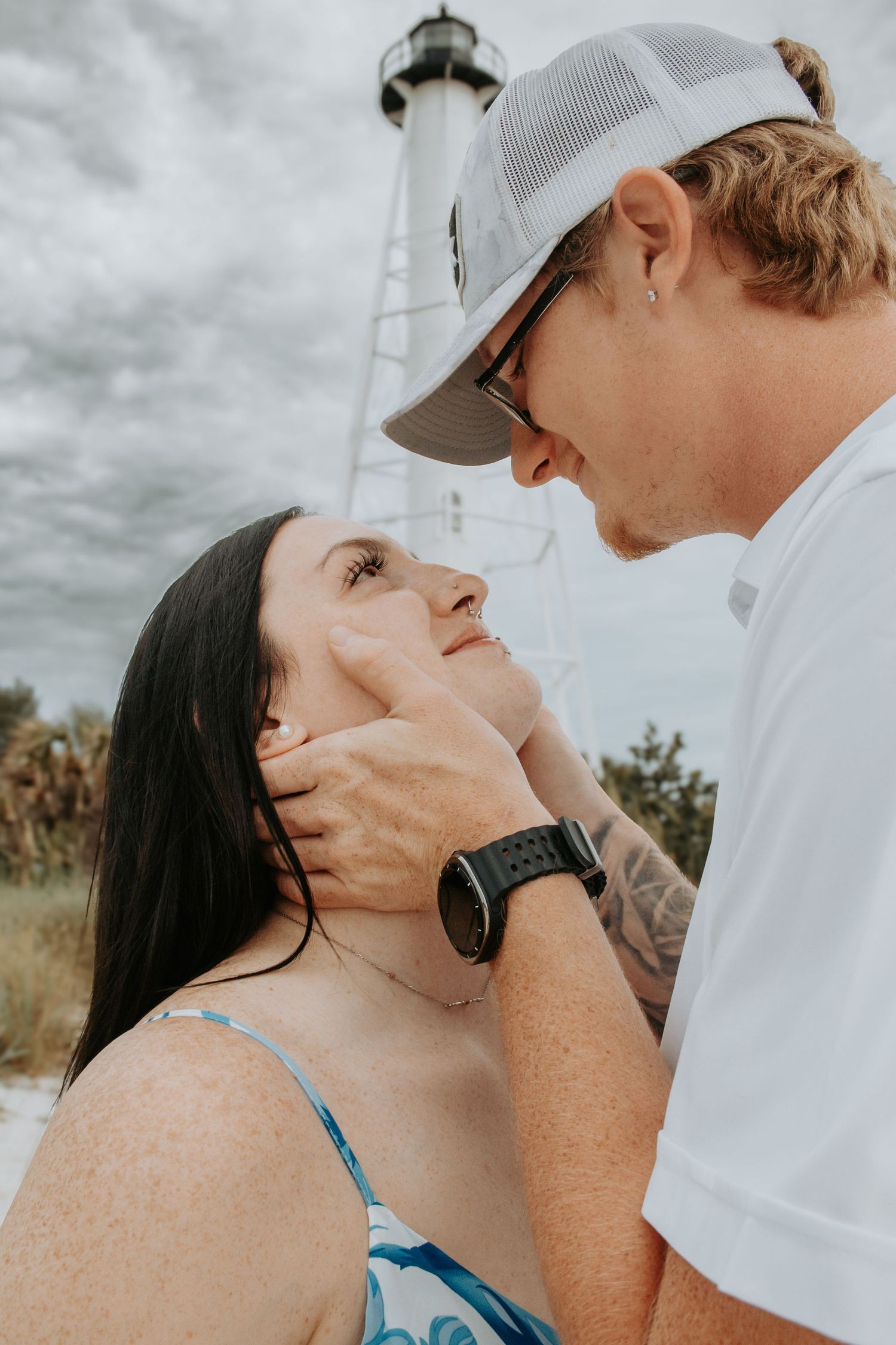 A couple shares a tender moment on the beach near a lighthouse, man gently holding womans face as she gazes up at him.