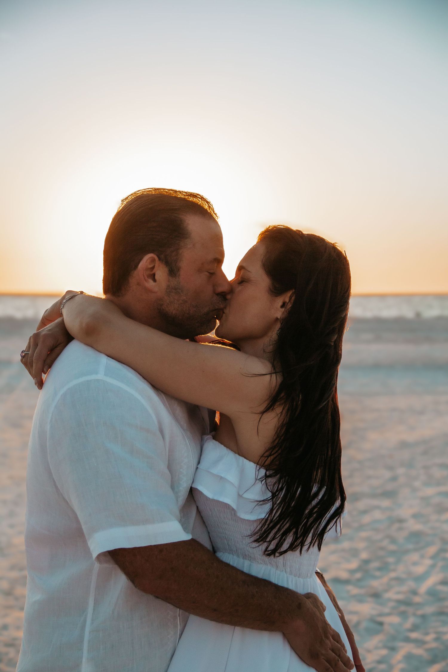 Couple sharing a romantic kiss on a sandy beach at sunset, both dressed in white outfits.