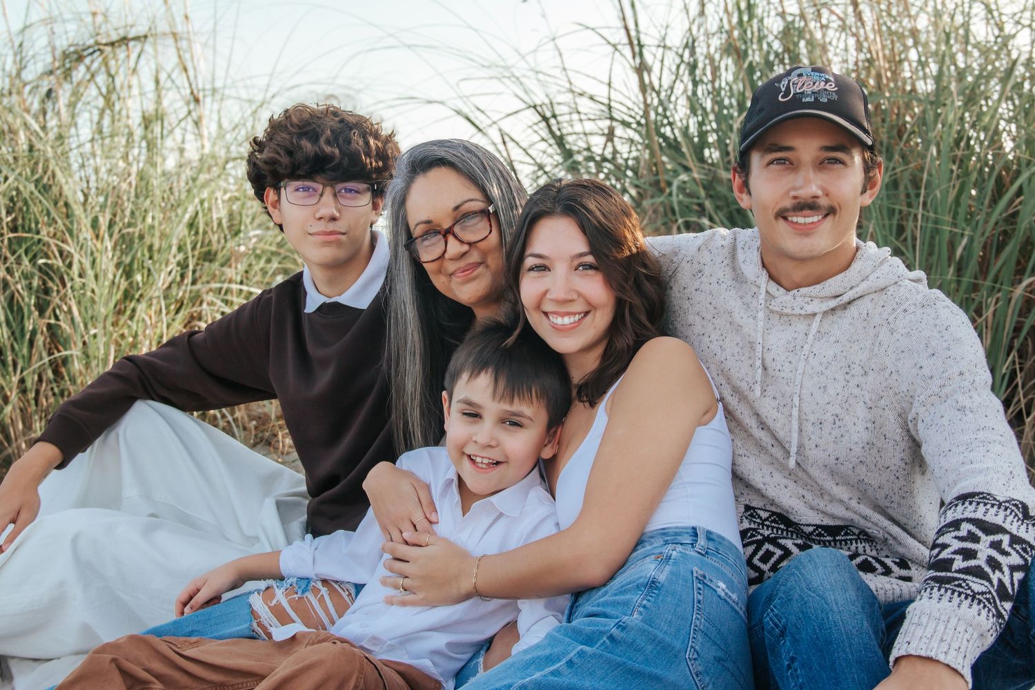 A multigenerational family of five smiles together outdoors near tall grass, enjoying a casual beach photo session.