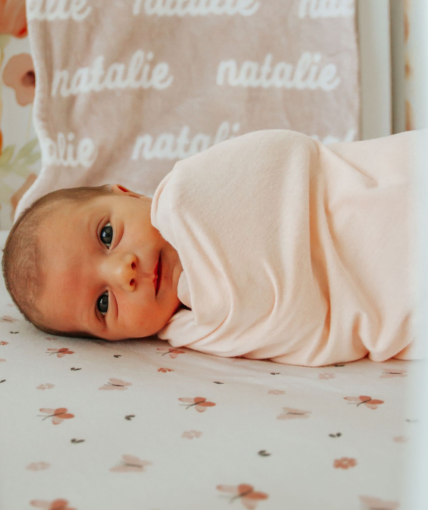 A newborn baby girl named Natalie swaddled in a pink blanket, lying on a floral crib sheet with a personalized name blanket.