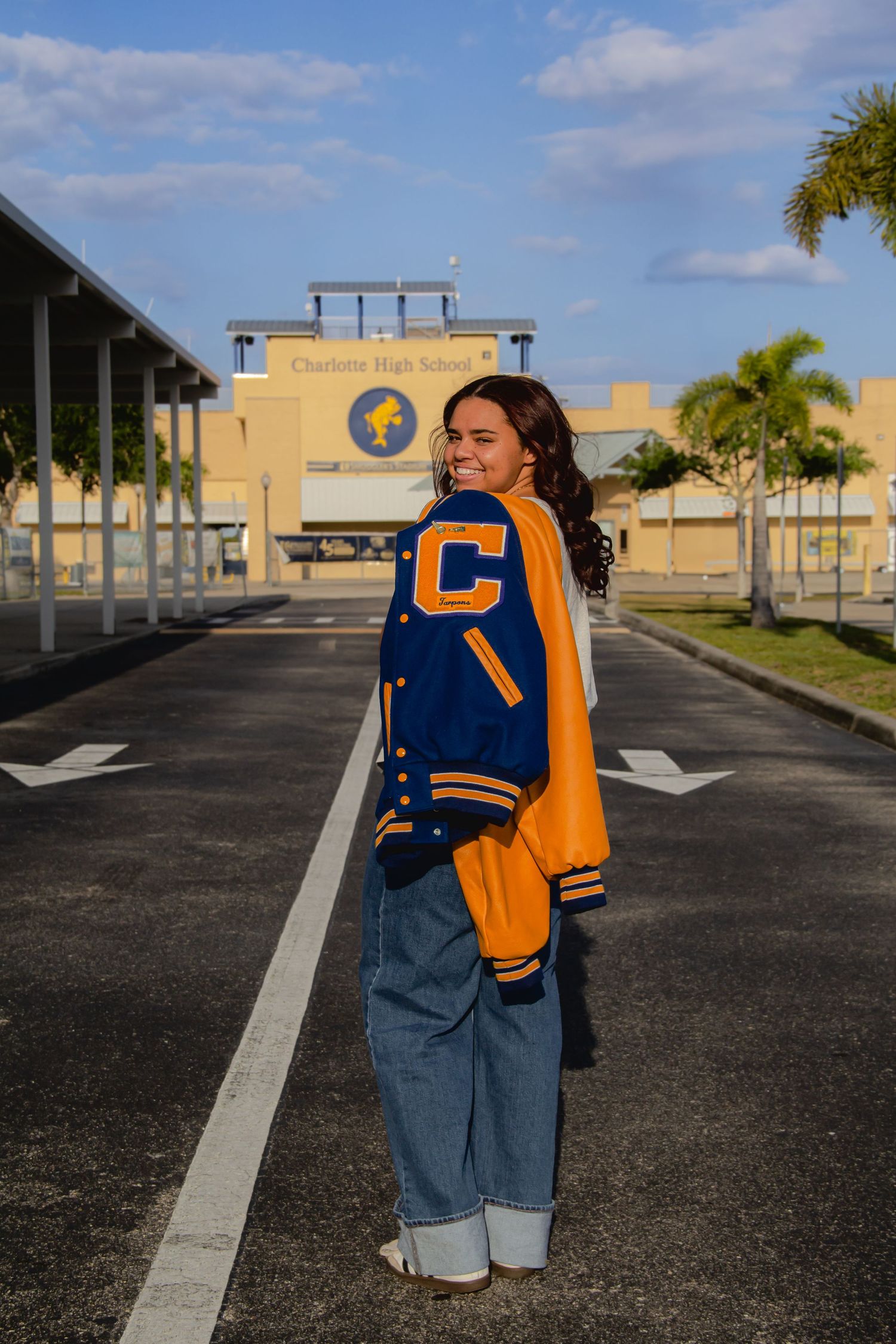 Smiling student in blue and gold varsity jacket poses in parking lot in front of Charlton High School building.
