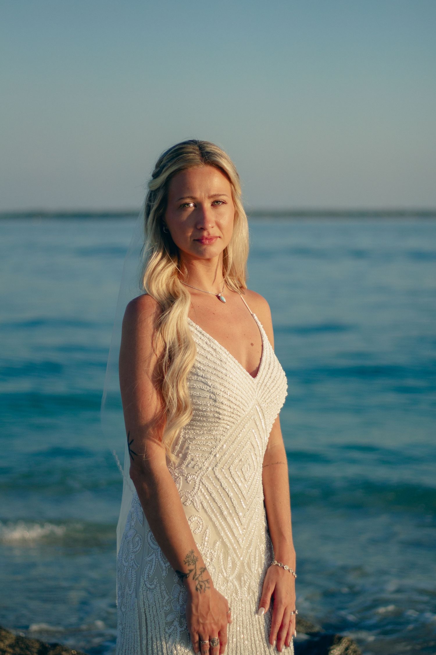 Blonde woman in white lace wedding dress standing on rocky beach with blue ocean backdrop at sunset.