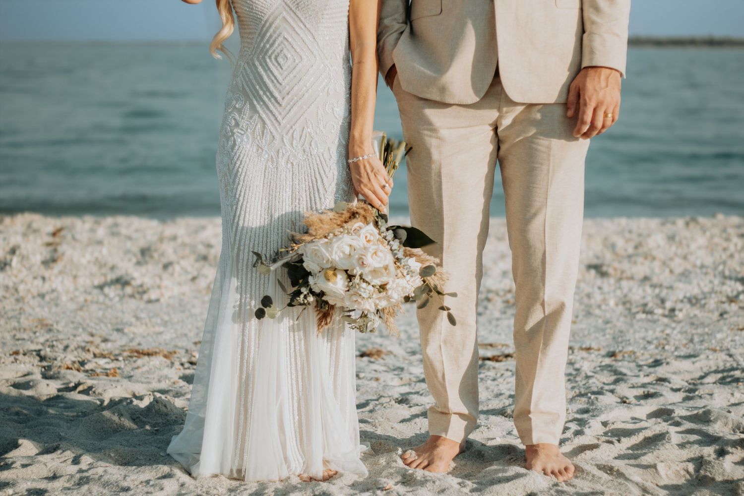 Barefoot bride and groom standing on sandy beach, bride holding white floral bouquet, groom in beige suit.