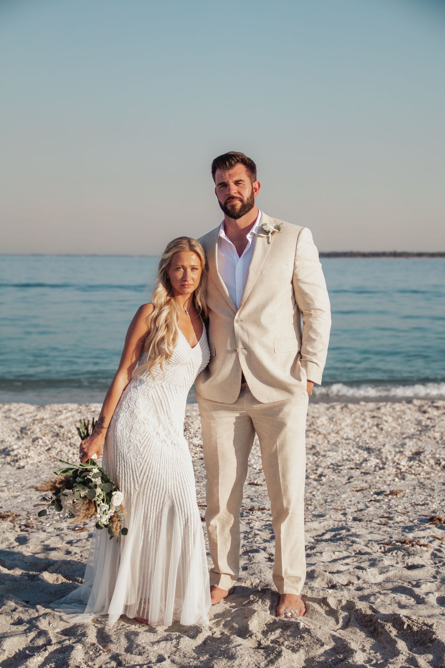 Bride and groom posing on a sandy beach, bride in white gown holding bouquet, groom in beige suit, ocean backdrop.