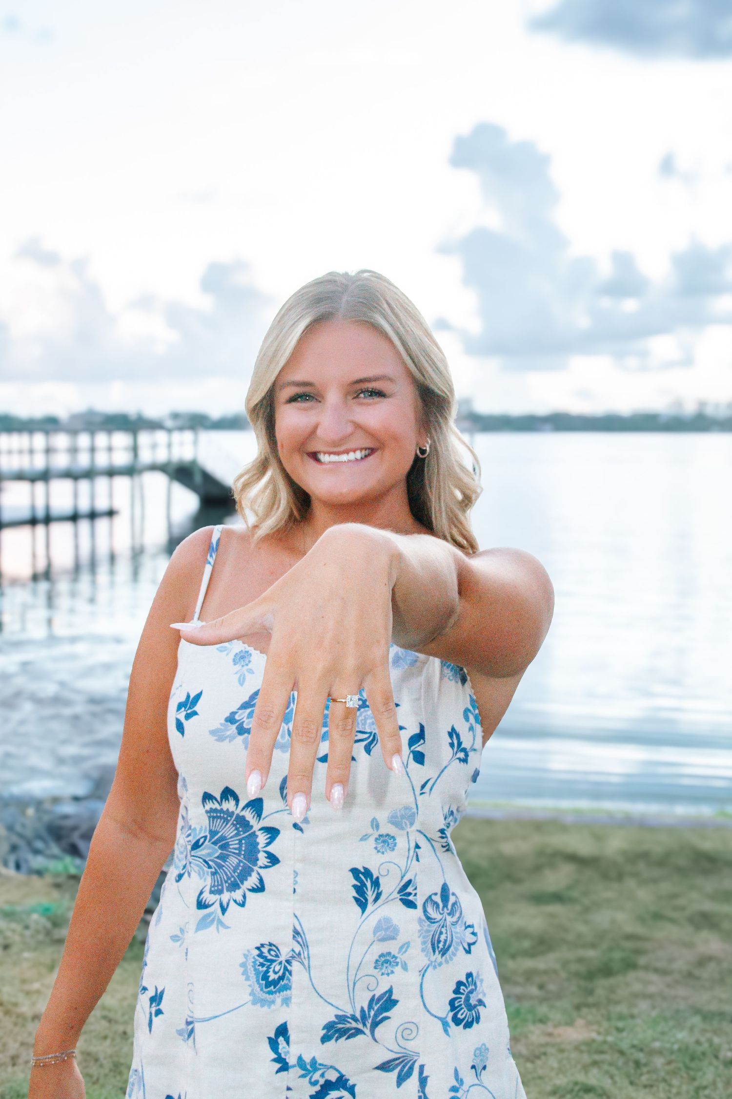 Smiling blonde woman in floral dress showing off engagement ring near waterfront dock.