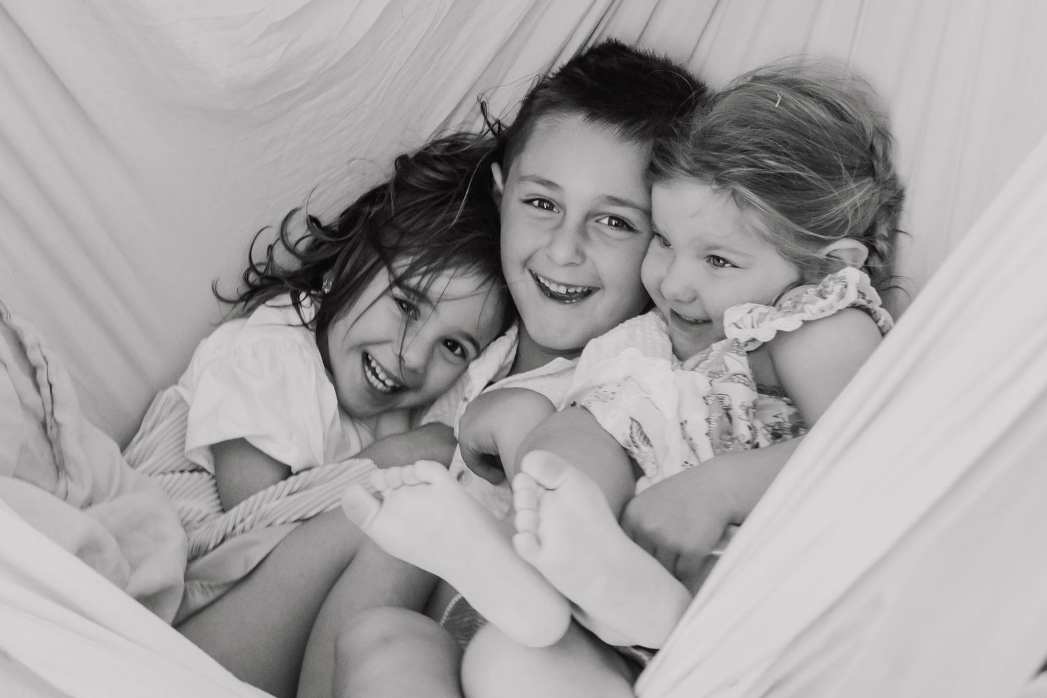 Three happy children cuddle together laughing in a cozy fabric hammock in a charming black and white photo.
