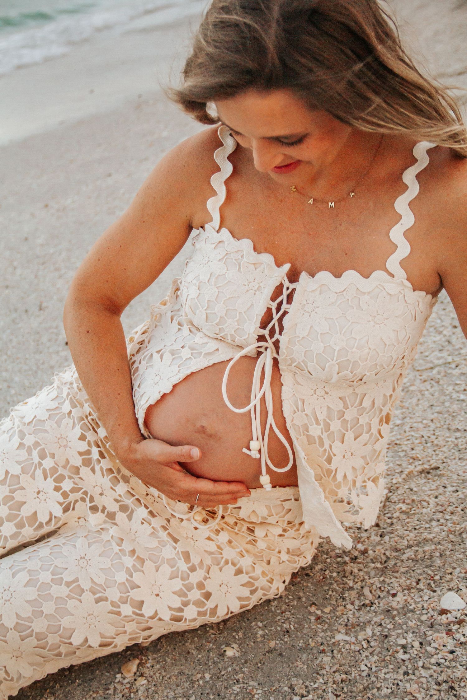 Pregnant woman in white lace crop top and skirt cradling baby bump on sandy beach.