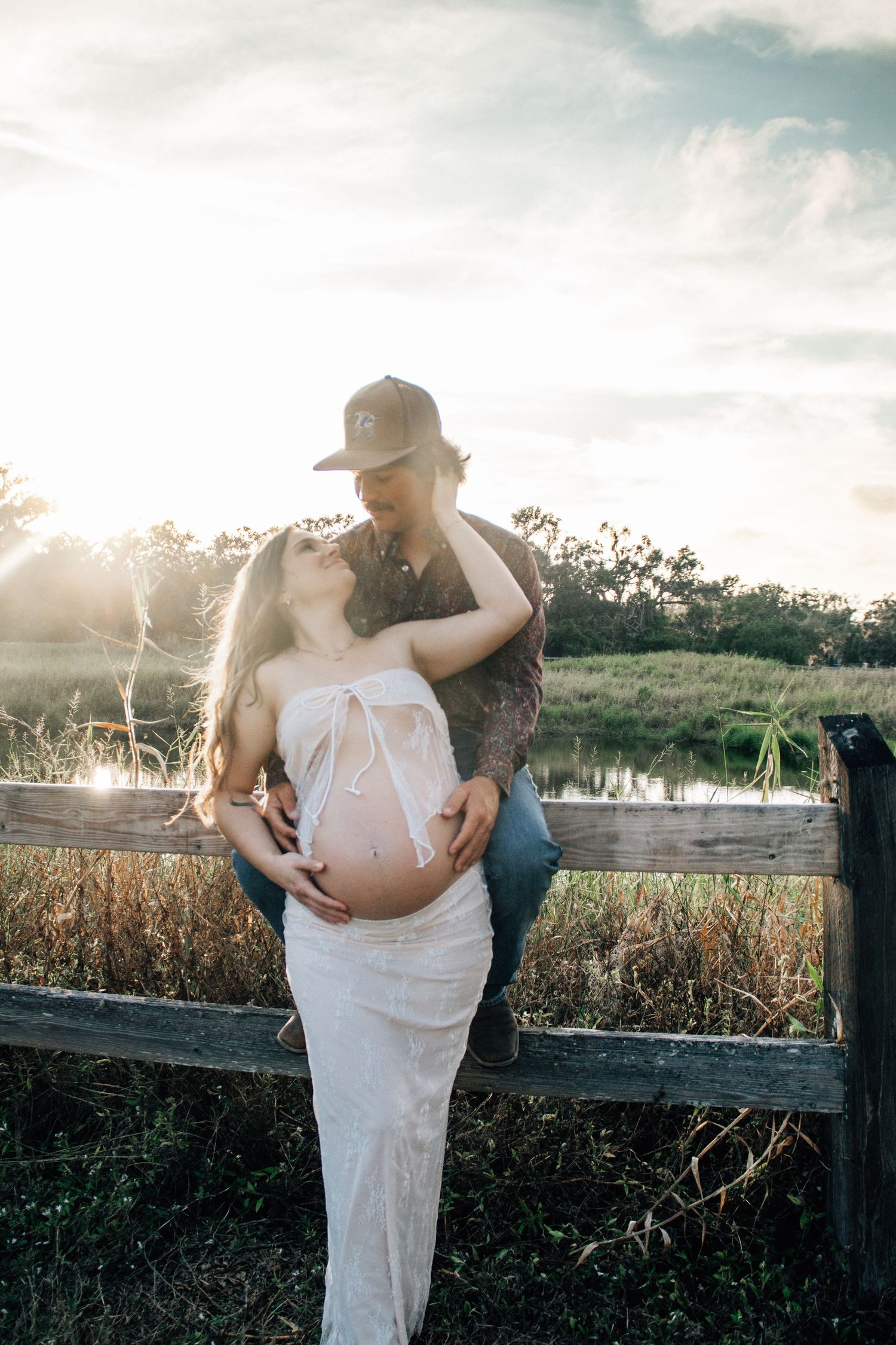Couple sharing a tender moment at sunset by a rustic fence, woman in white dress cradling baby bump during maternity photoshoot.