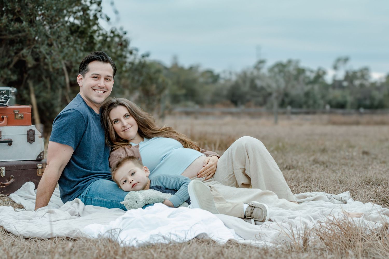 A happy family of three relaxing on a blanket in a field, with a pregnant mother, father, and young child.