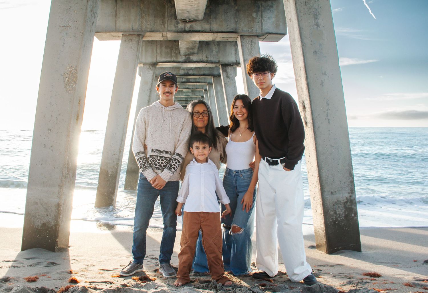 Family of five posing under a beach pier on sandy shore with ocean waves in the background.