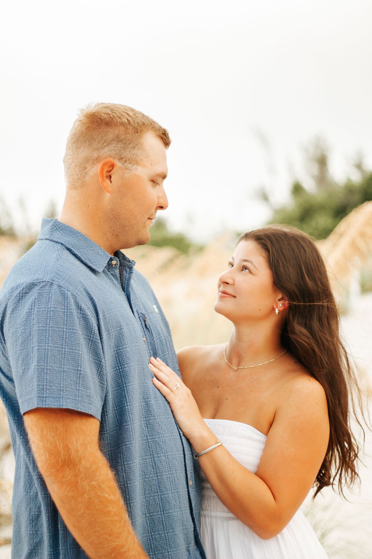 A couple shares a romantic moment on a beach, woman in white dress gazing up at man in blue shirt.