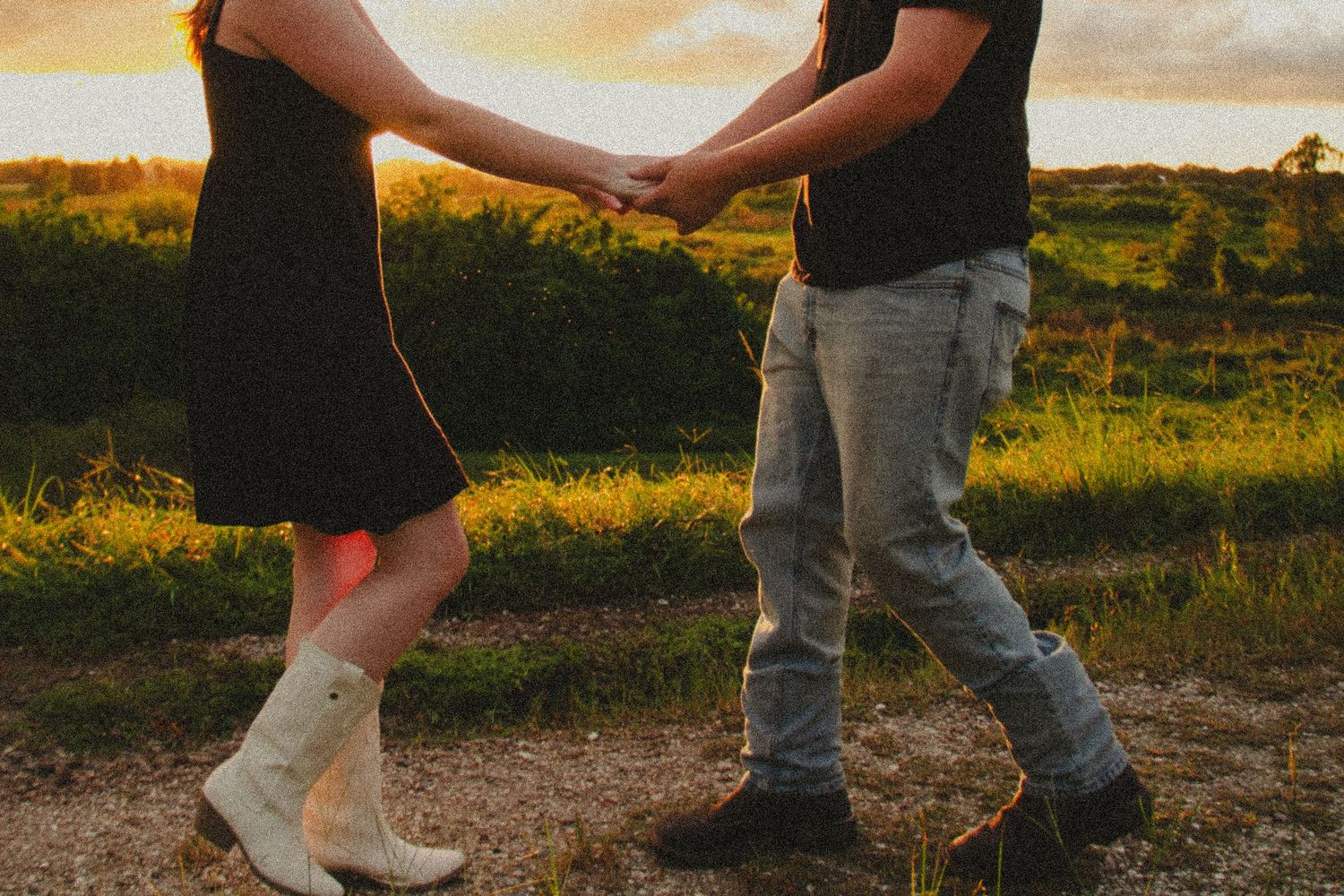 Couple holding hands dancing at sunset in a field, woman wearing white cowboy boots and black dress.