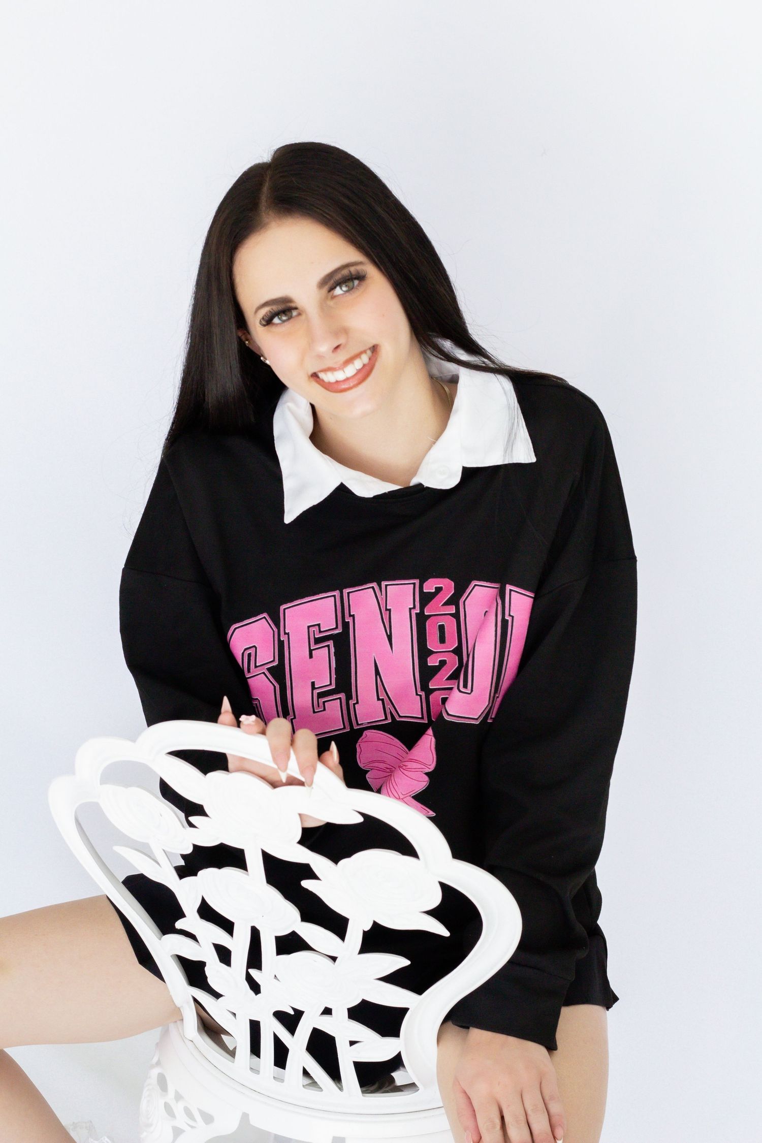 Smiling young woman with dark hair wearing a black Senior 2024 sweatshirt over white collar shirt, seated on white chair.