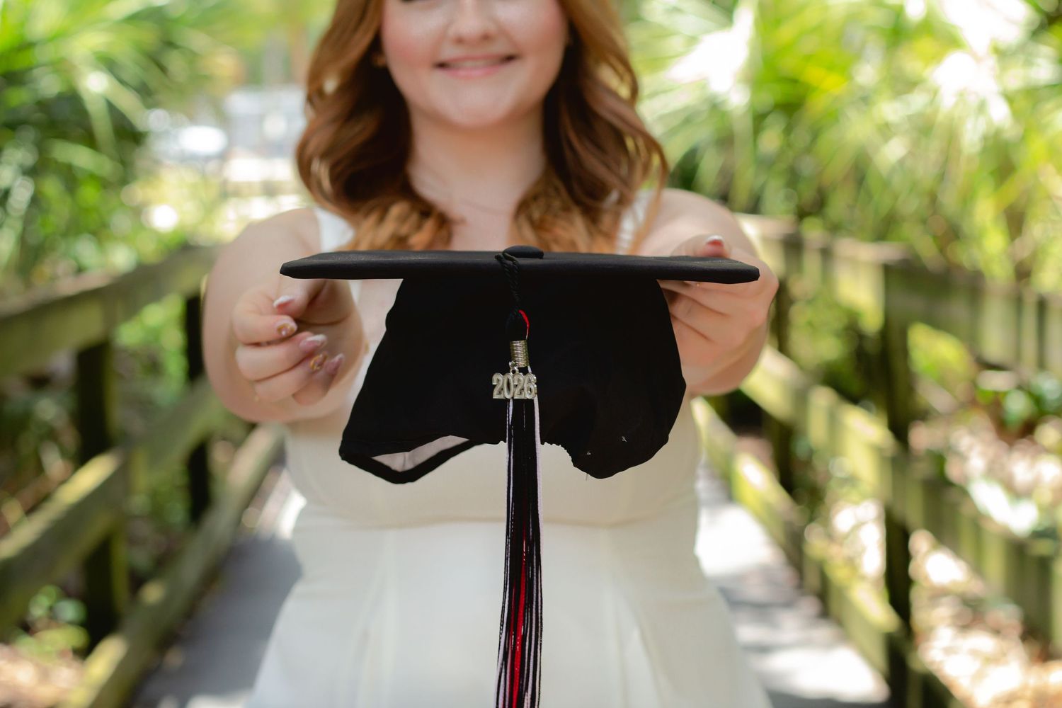 Young woman in white dress holding a black graduation cap with red tassel outdoors on a wooden bridge.