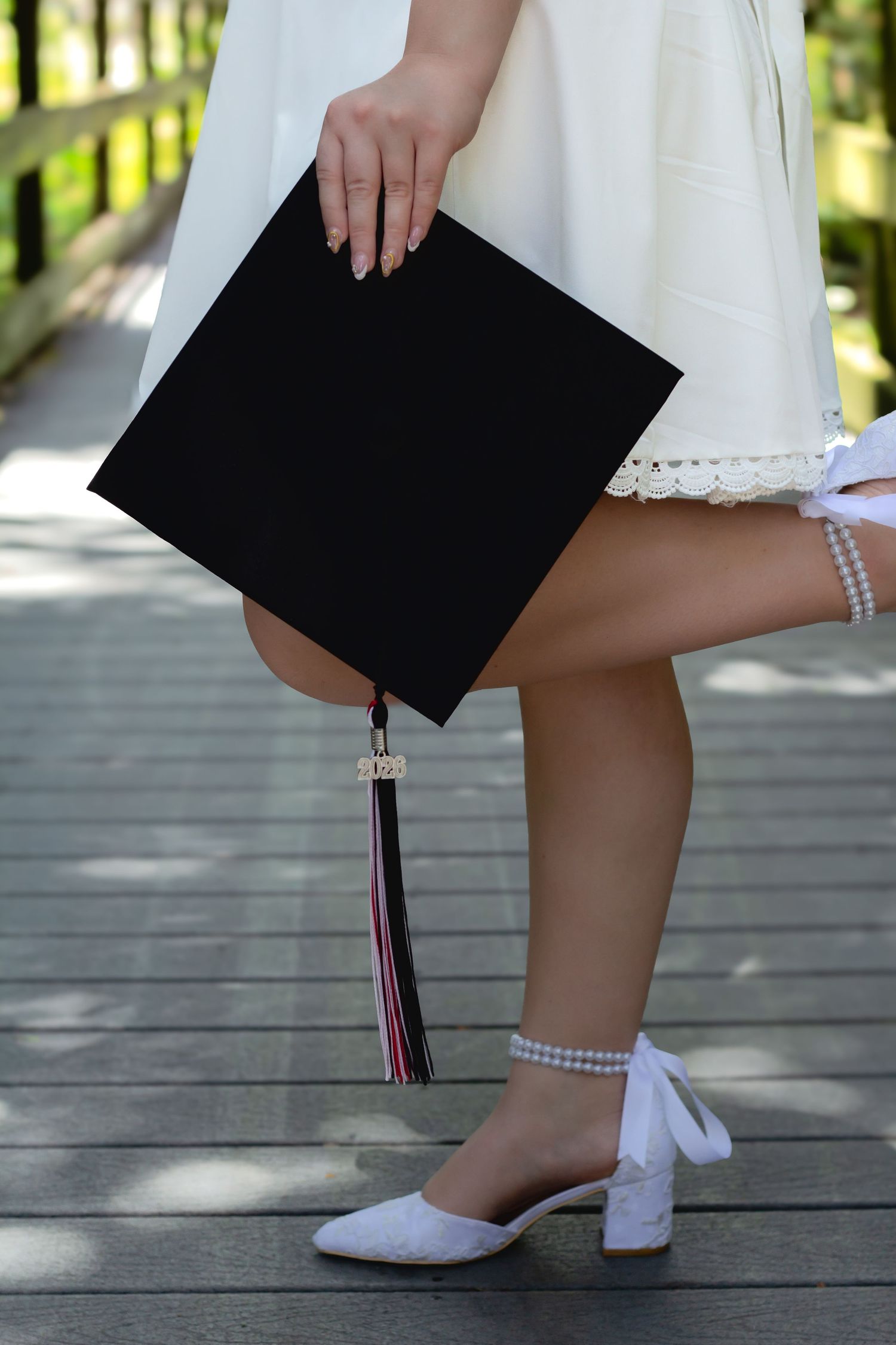 Graduate in white dress holding black mortarboard cap with tassel, wearing white heels on wooden bridge.