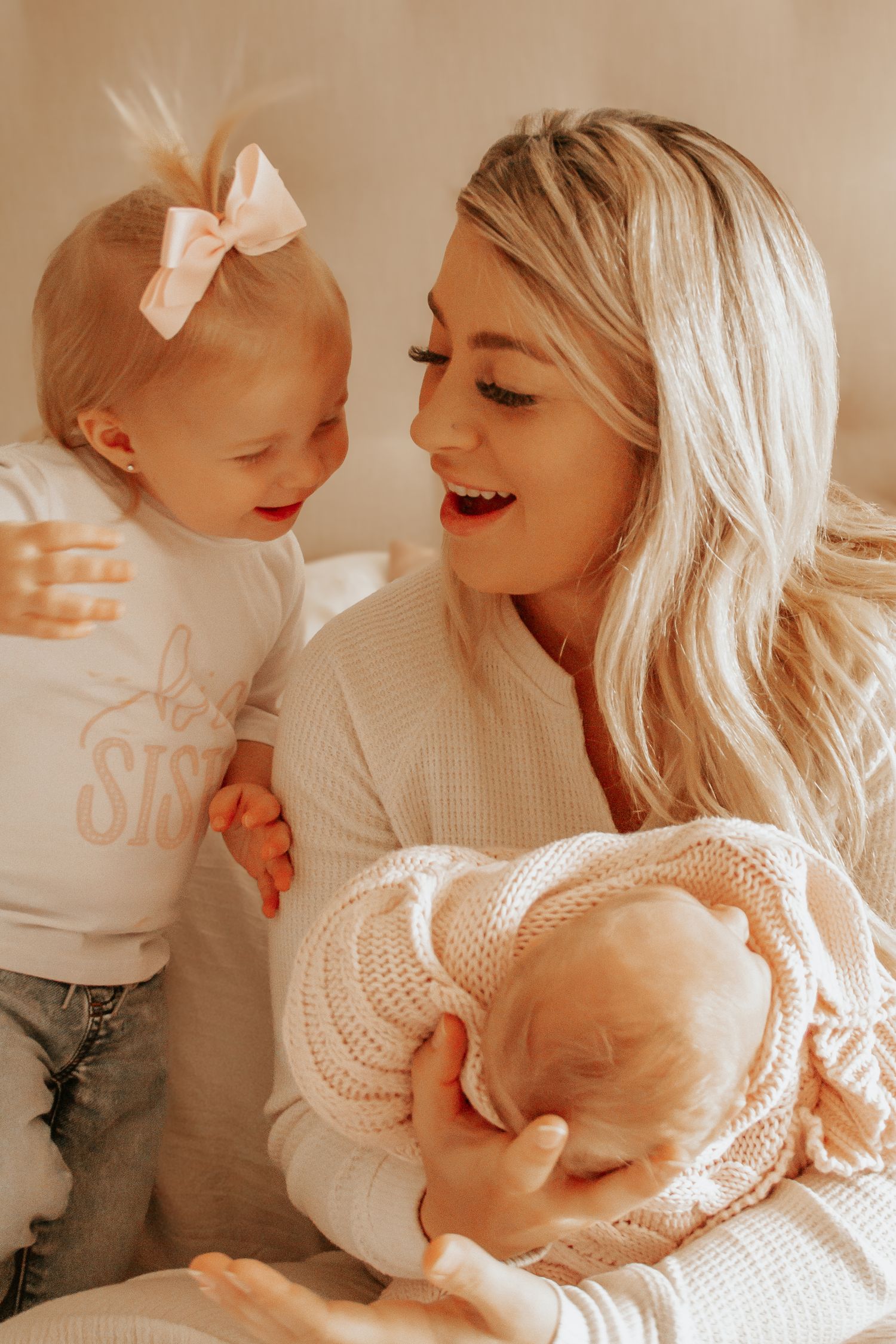 Smiling blonde mother holds newborn baby while toddler girl in big sister shirt looks on adoringly.