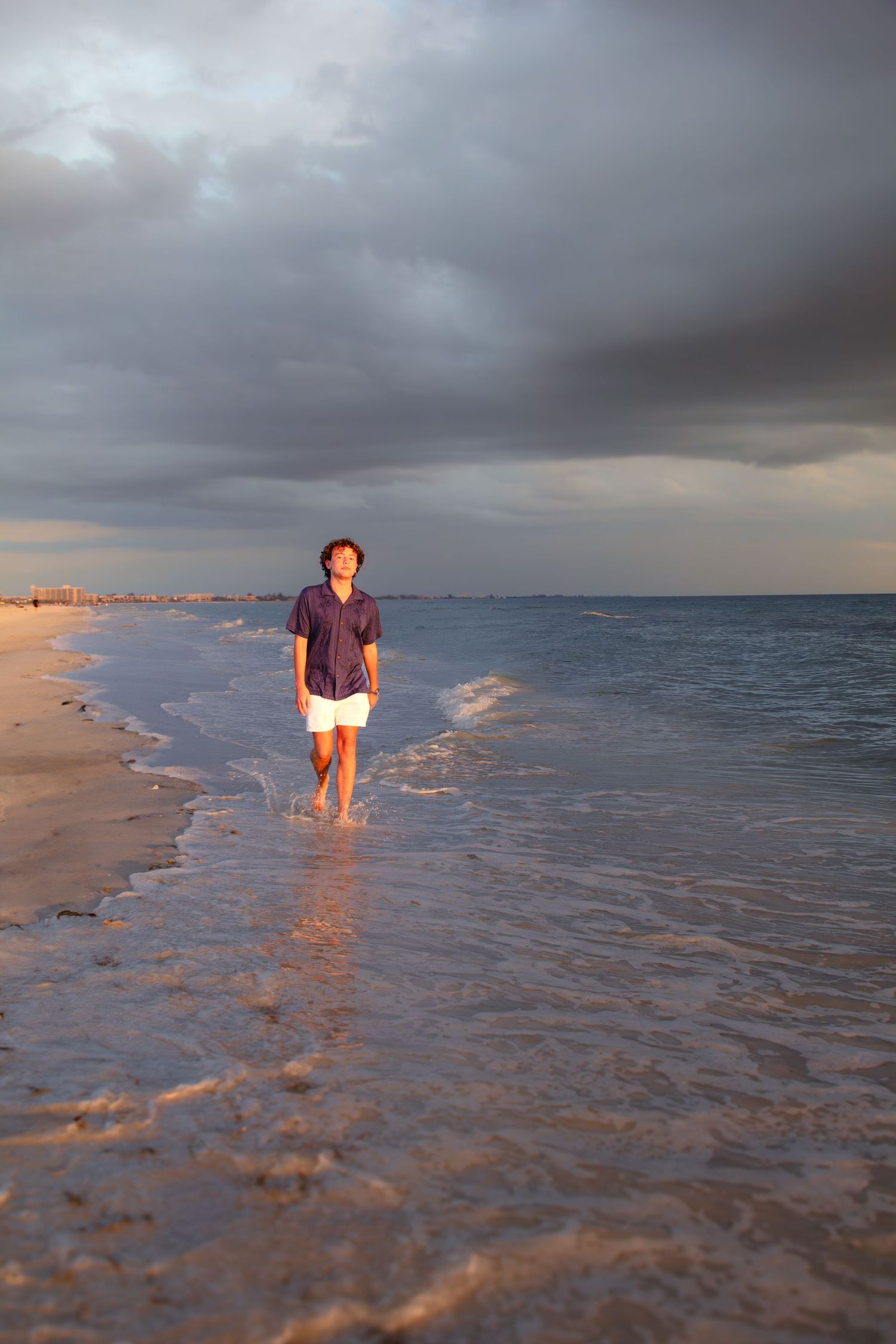 Man walking along shoreline at sunset with dramatic storm clouds over ocean, wearing purple shirt and white shorts.