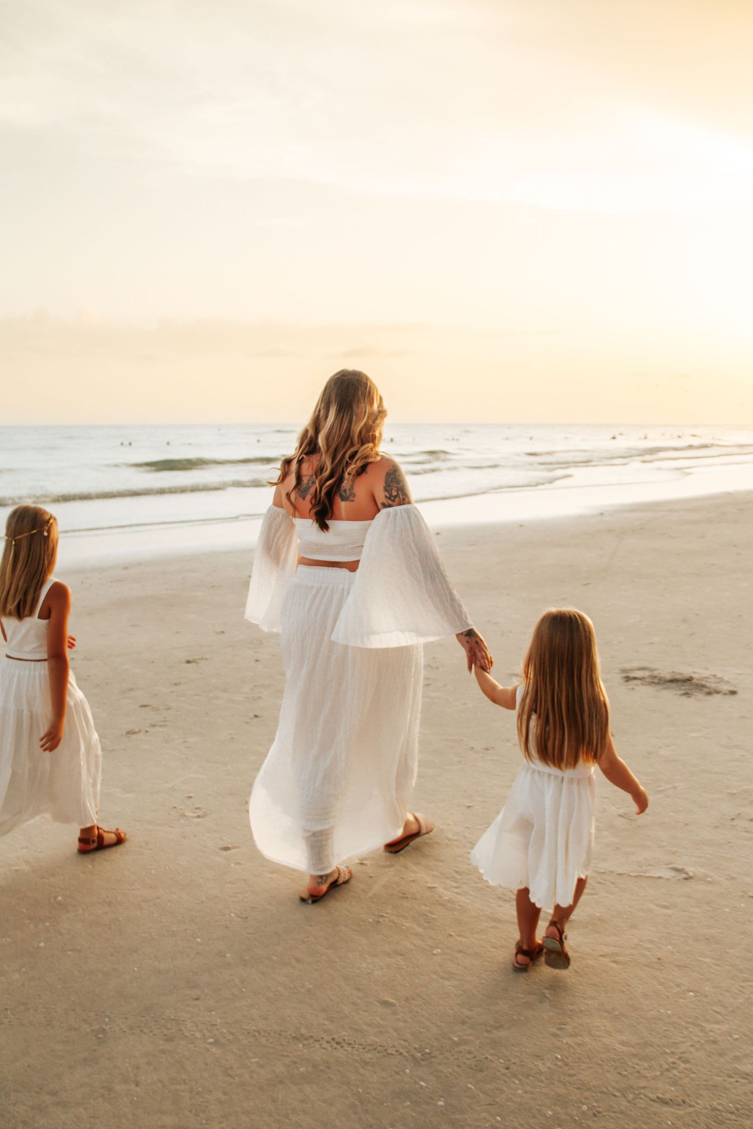 A mother and two daughters in white dresses walk hand-in-hand on a sandy beach at sunset.