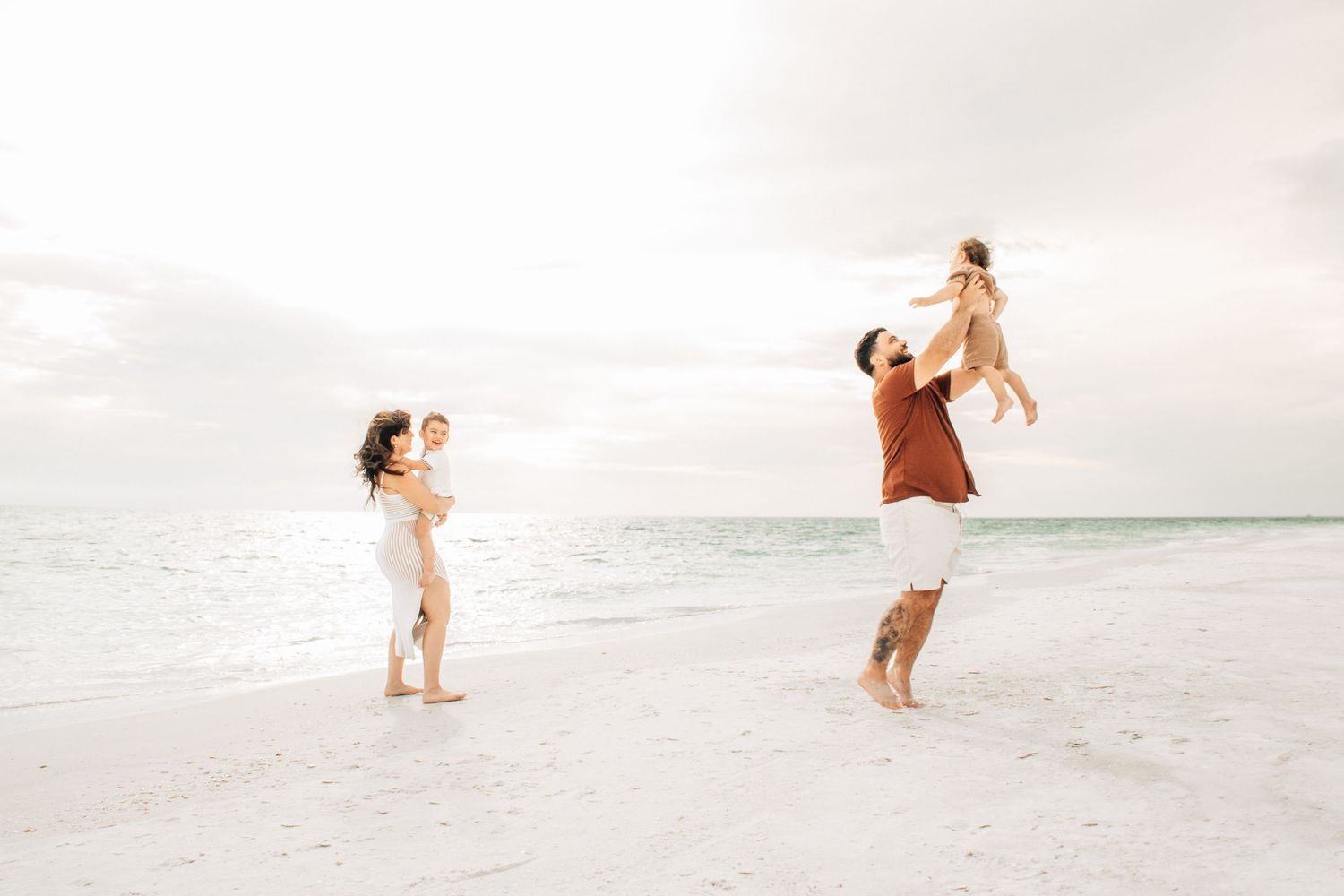Family enjoying a sunny beach day, parents playfully lifting children on white sand shore with calm ocean waves.