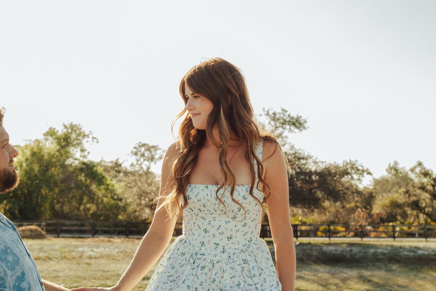 Young woman in floral sundress with long wavy hair standing outdoors in warm sunlight near trees.
