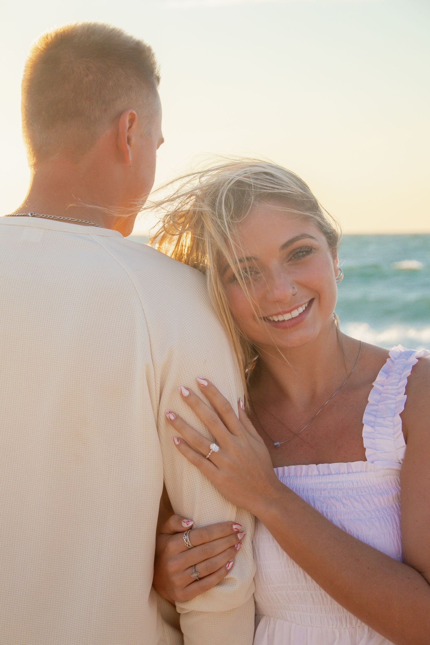 Happy woman in white dress hugging man on beach at sunset, showing engagement ring, ocean in background.