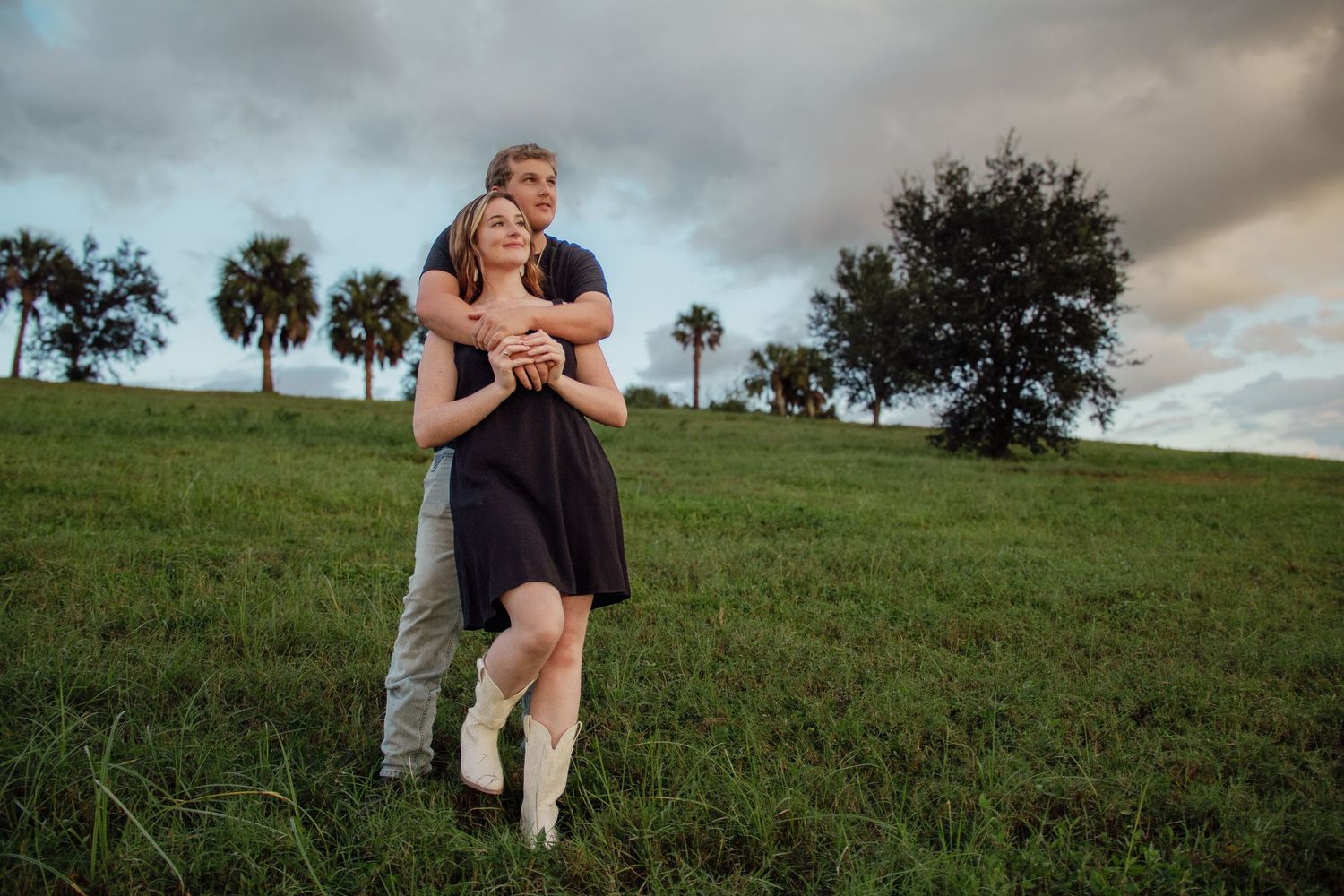 Couple embracing on lush green hill with palm trees and dramatic cloudy sky in outdoor engagement photo.