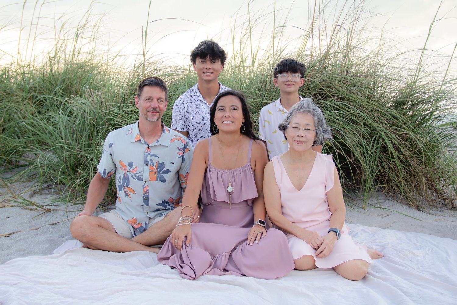 Family of five smiling together on a sandy beach with tall grass in the background.