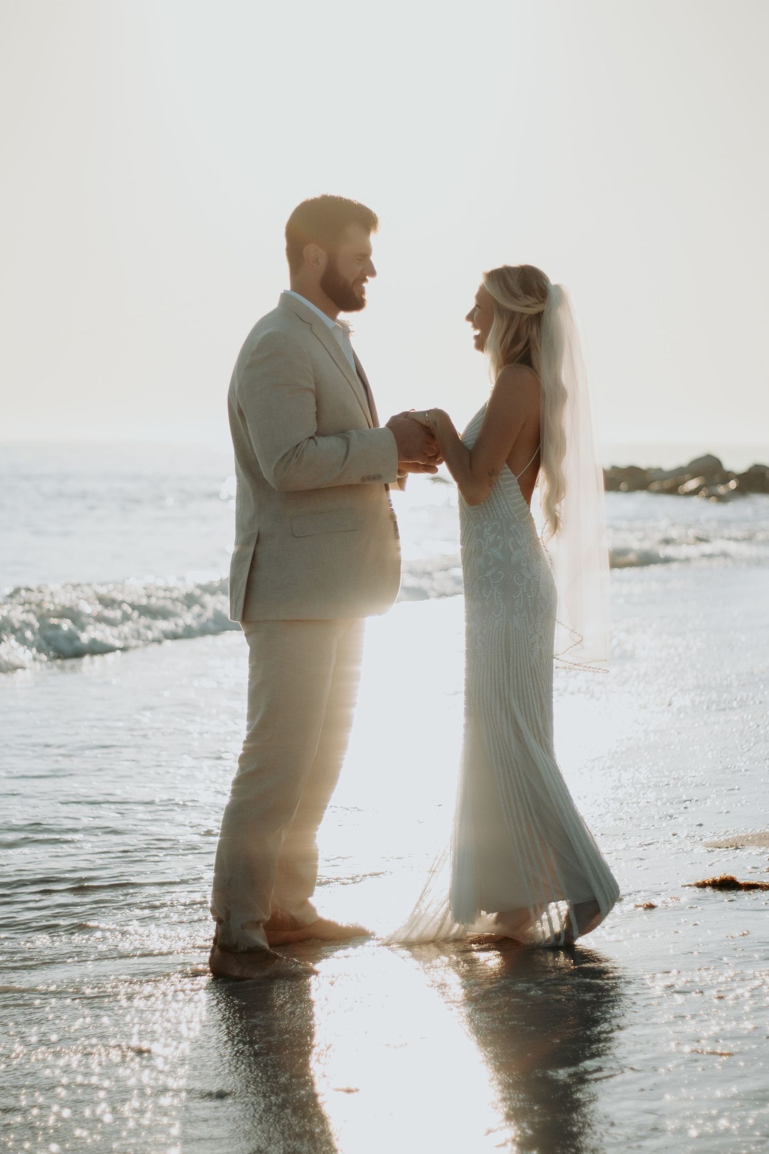 Bride and groom holding hands on a sunlit beach, waves rolling in behind them during a romantic outdoor wedding ceremony.