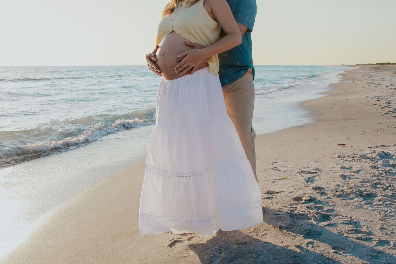 Pregnant couple embracing on a sandy beach at sunset, woman in white maxi skirt cradling baby bump near ocean waves.