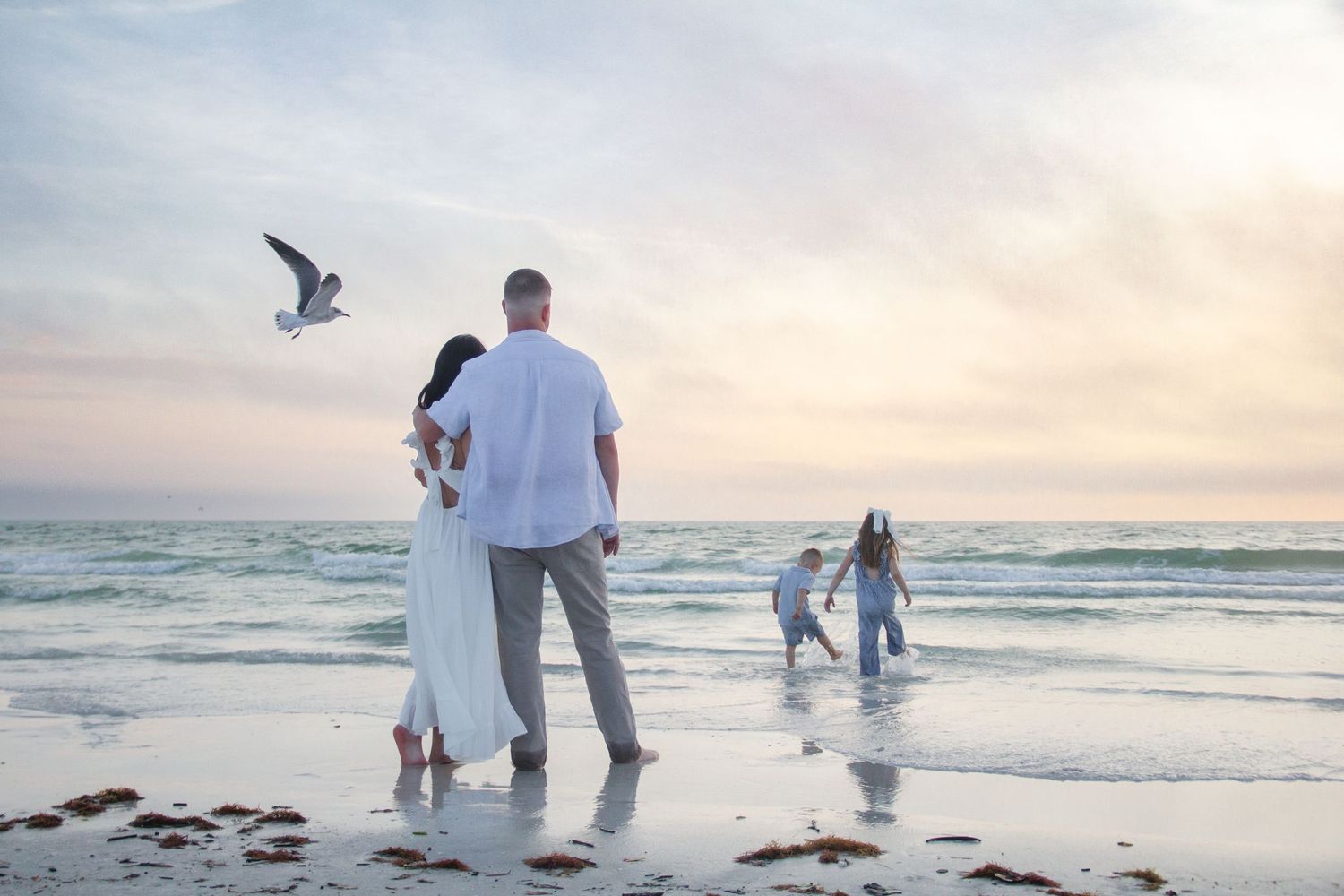 Family enjoying a serene beach sunset, couple embracing while children play in the waves with a seagull flying overhead.