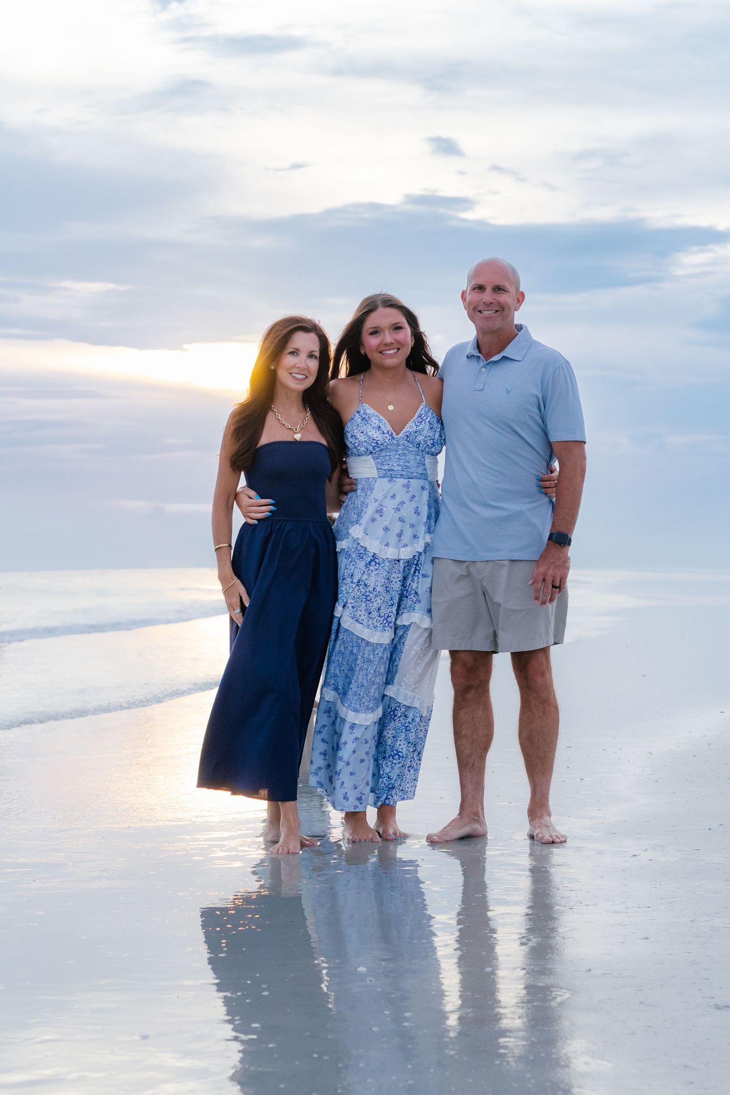 Family of three smiling together on a beach at sunset, wearing blue outfits, standing on wet sand with ocean waves.