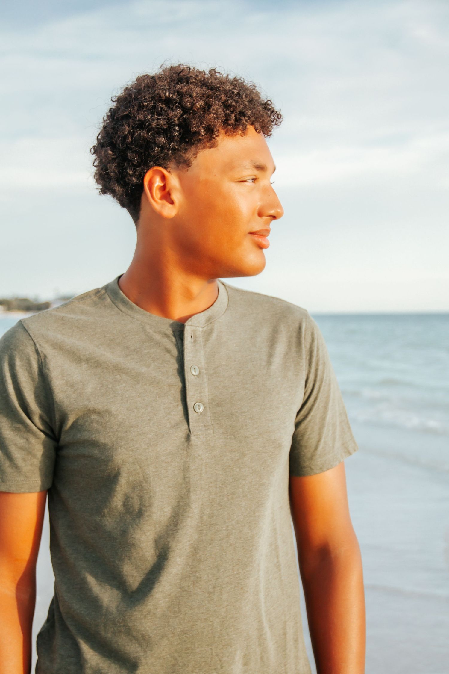 Young man with curly hair wearing a gray henley shirt standing on a beach with ocean waves in the background.