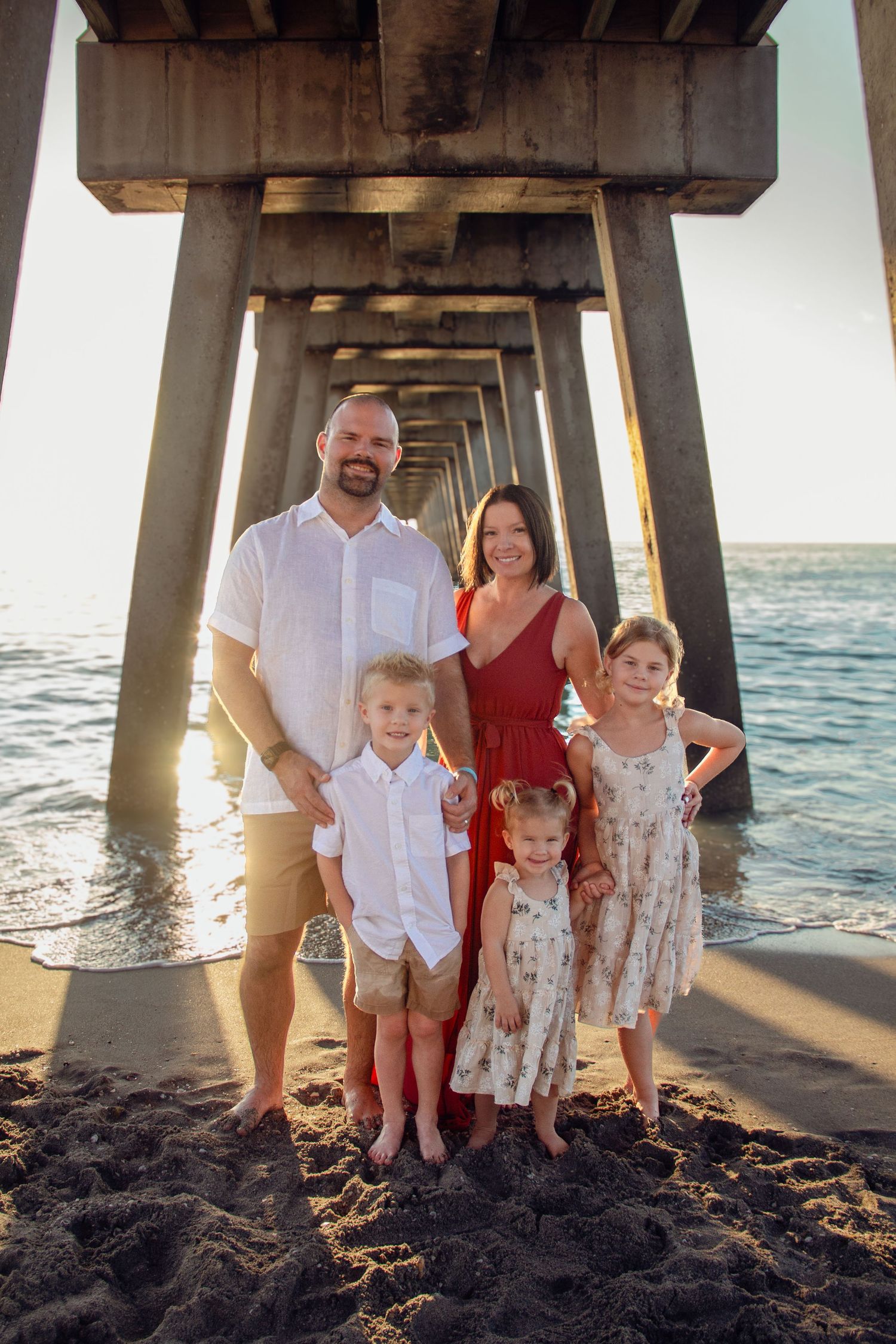 Family poses under a wooden pier at sunset on a sandy beach with ocean waves in the background.