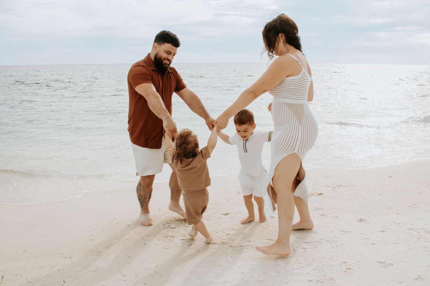 Family of four holding hands and dancing on a sandy beach with calm ocean waves in the background.