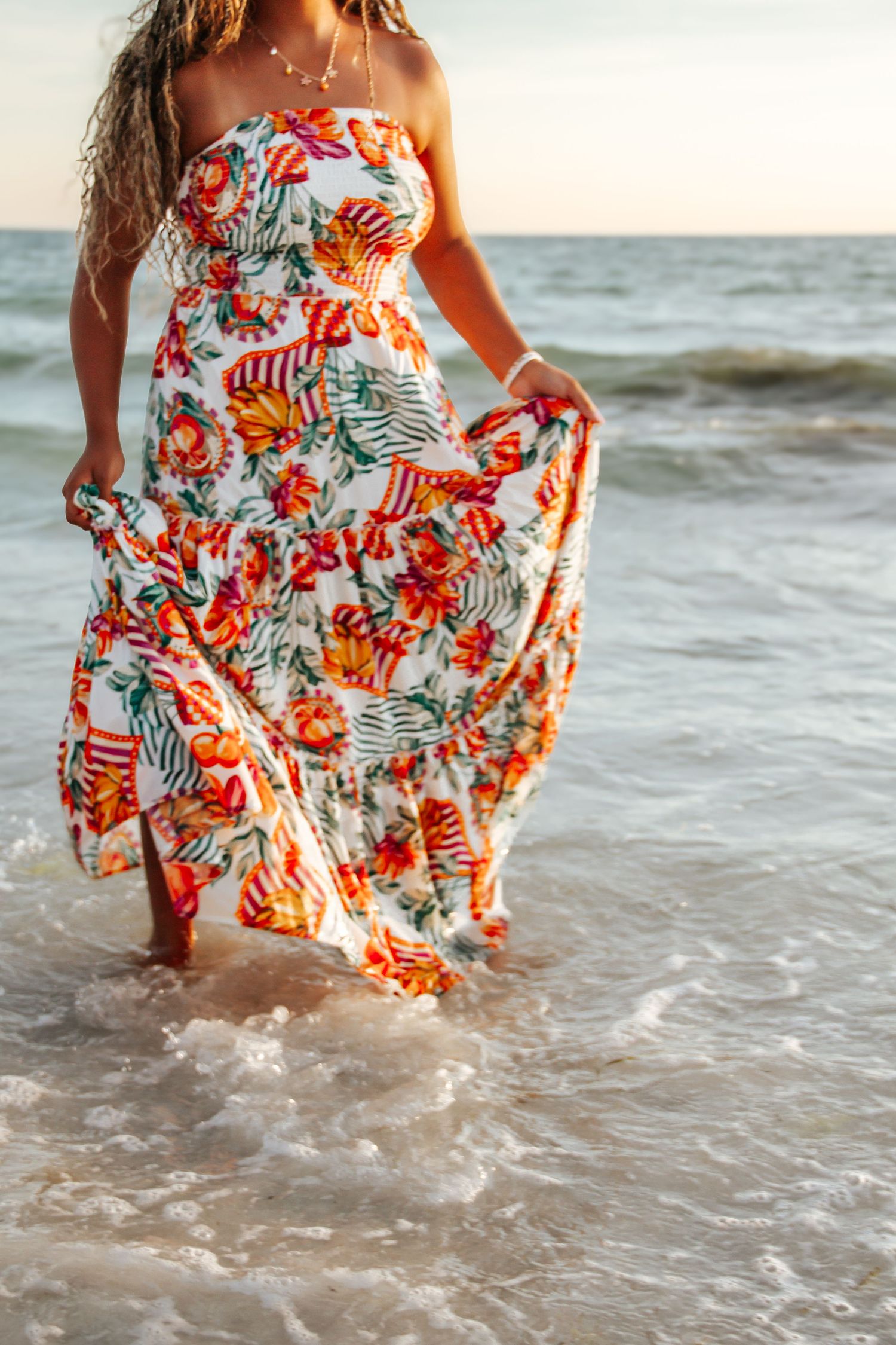Woman in floral maxi dress wading in ocean waves at sunset on a beach.