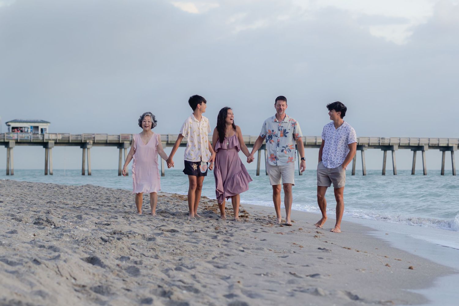 Family of five holding hands walking on a sandy beach near a pier at sunset.