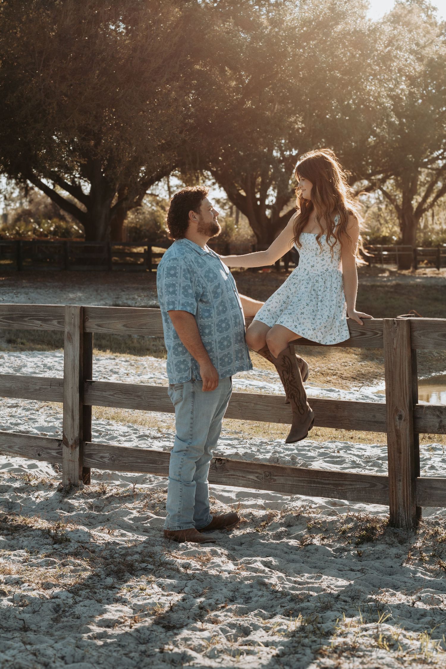 Man and woman in floral dress sitting on wooden fence sharing a romantic moment in warm sunset light.